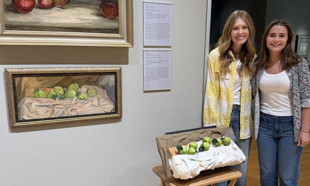 Two young women stand in an art gallery with paintings on the walls. One painting shows a groups of seven green apples arranged in a messy line on a rumpled white cloth. Next to the women, on a small folding table, is a 3-D replica of the painting.