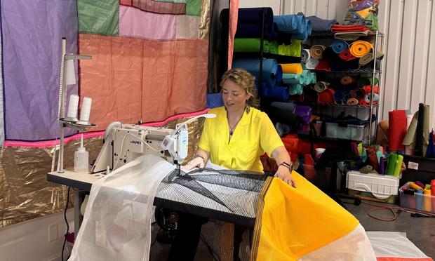 A woman (Rachel Hayes) sitting at a sewing machine in a textile studio. She’s surrounded by colorful fabric and wears a bright yellow top.