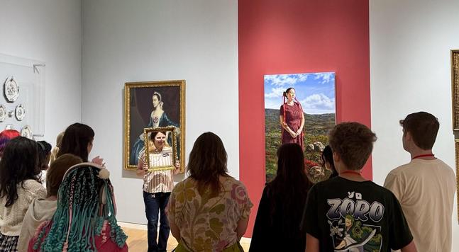 A group of people in a museum gallery, on a tour. In front of them, a tour guide holds up an empty frame around her face, smiling.