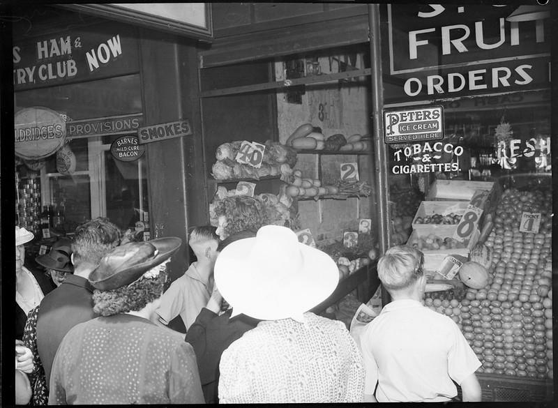 Black and white photograph of people crowding outside of a fruit market.