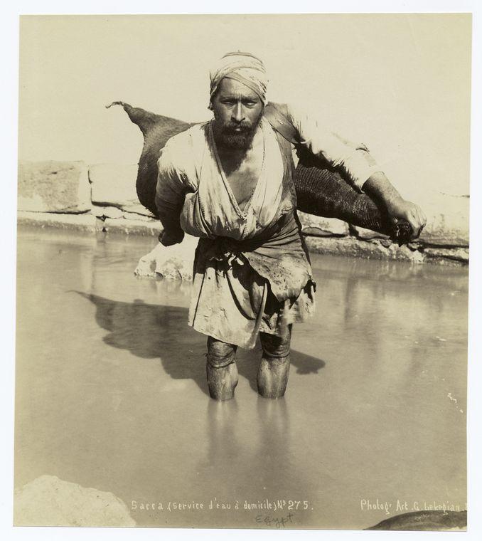 A water delivery man in a turban carries a goatskin of water through a watery area. He is staring directly at the camera.
