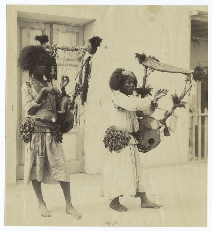 Two street musicians playing instruments with strings and a large resonator which rests on their hips