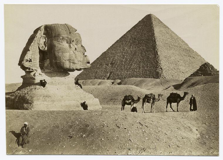 Sepiatone photograph of the Sphinx in front of a large and small pyramid. There are people on and with camels in the foreground.