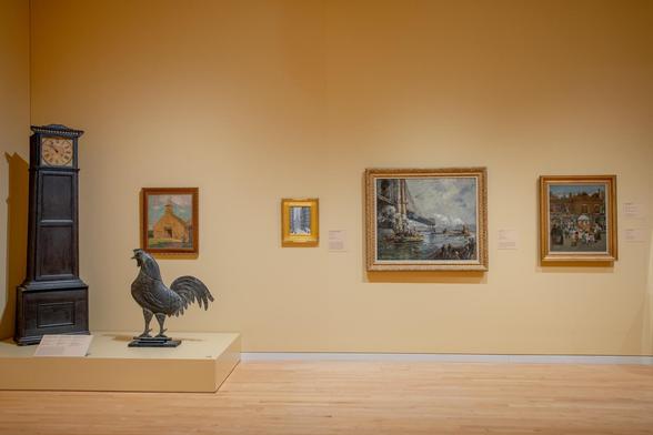 An installation photo of the museum’s Marilyn Overstreet Nalley South Gallery, showing a tall case clock, a large rooster-shaped metal weathervane and several paintings hung on the butter-yellow wall.