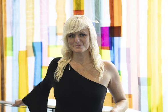 Lily Herne, Museum Shop associate at the Georgia Museum of Art, stands in front of a colorful translucent textile piece hung on the windows. She wears a one-shouldered black top and her blond hair falls below her shoulders.