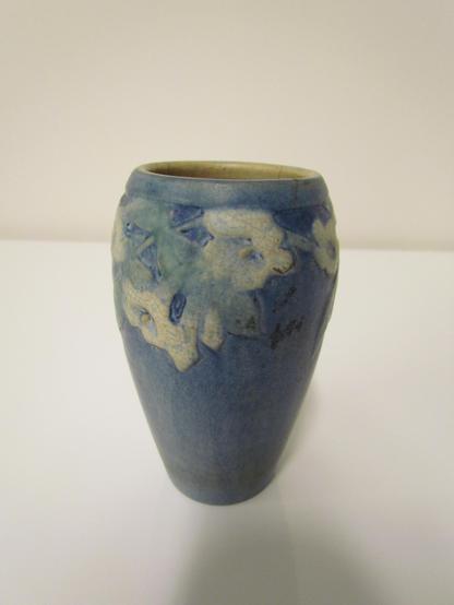 A Newcomb Pottery vase, with a blue glazed exterior (more matte than shiny) painted with slightly raised images of white trumpet flowers, a la morning glories or moonflowers, as well as their vines and leaves. The flowers ring the top half of the vase, which is wider at the top than at the bottom but otherwise cylindrical.